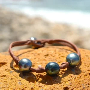 Brown leather bracelet with three central 9.5 mm Tahitian pearls, knots and two closure pearls displayed on a rock by the sea