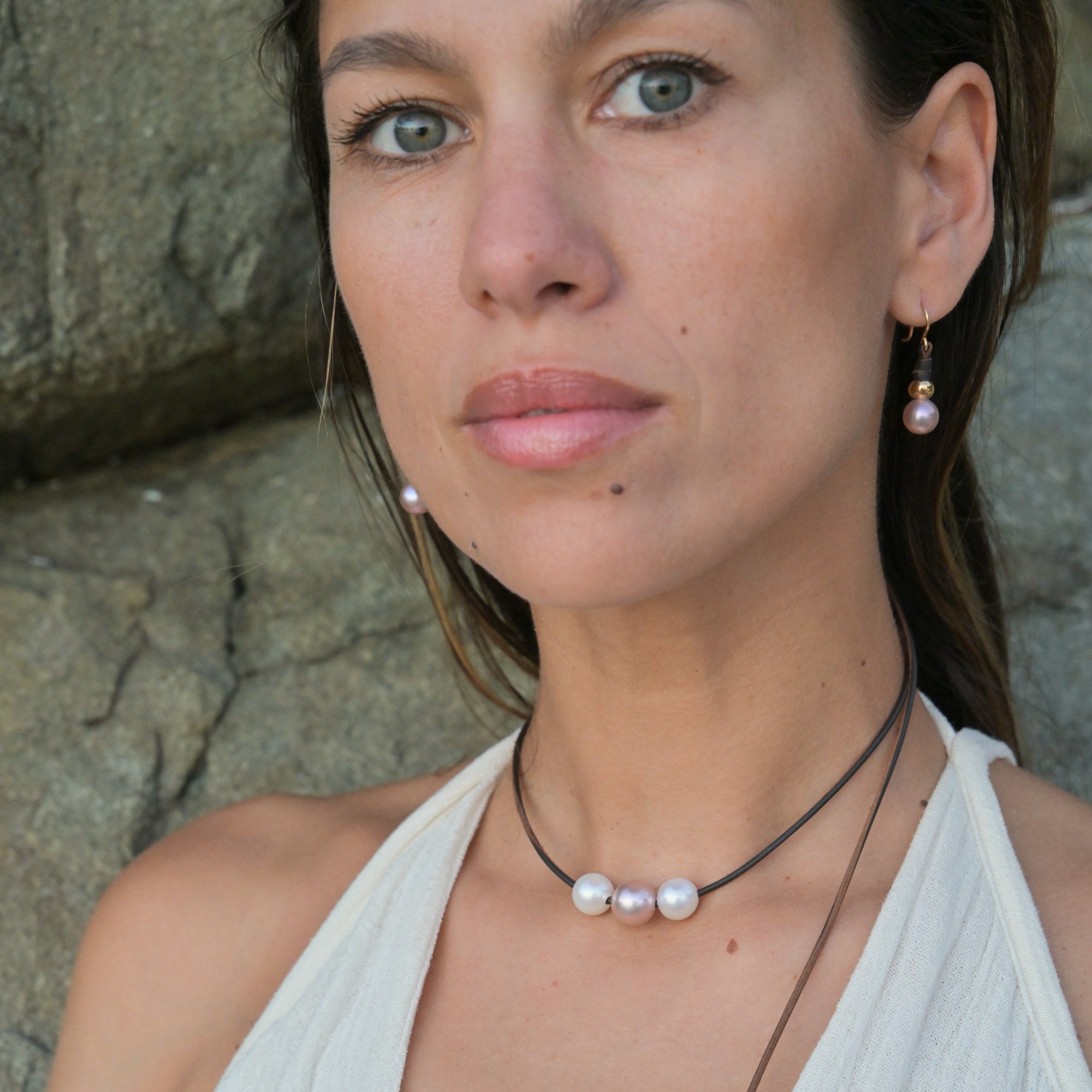 Boucles d’oreilles en cuir, perles roses, billes dorées et crochets en or, vue portée devant les rochers, portrait de face.