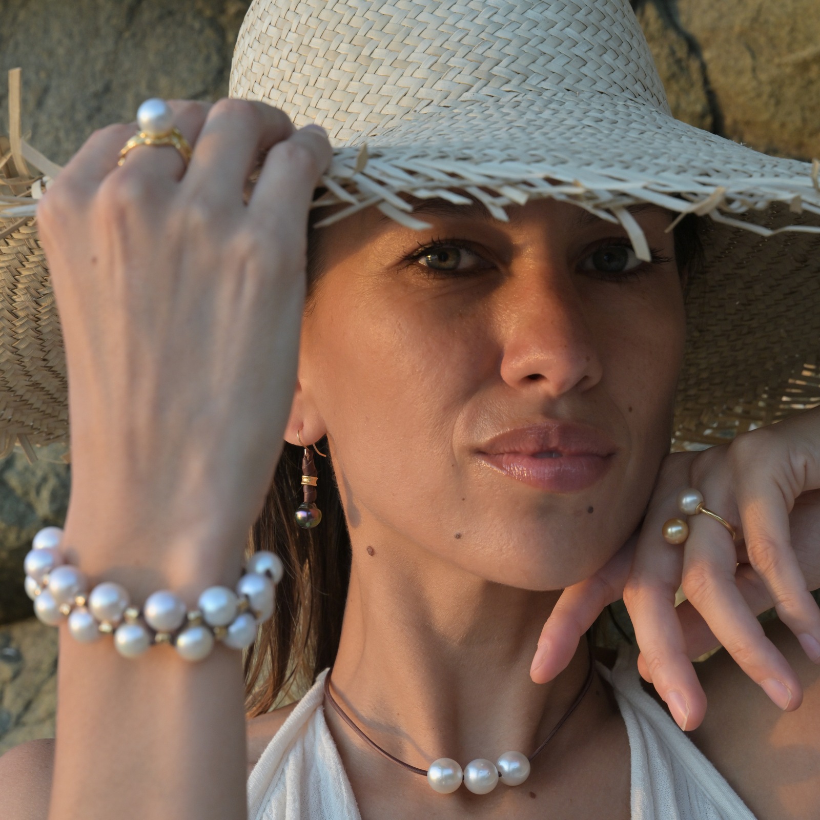 Boucles en cuir, perles vertes de Tahiti et crochets en or, vue portée avec un chapeau de paille, la main près du visage.