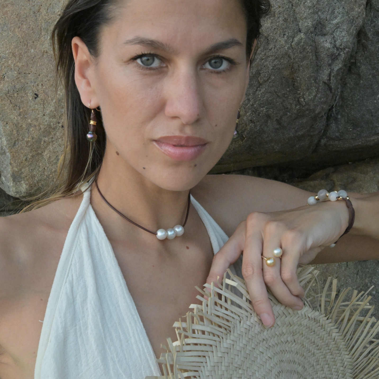 Boucles en cuir, perles vertes de Tahiti et crochets en or, vue portée devant les rochers, avec un chapeau tenu à la main.