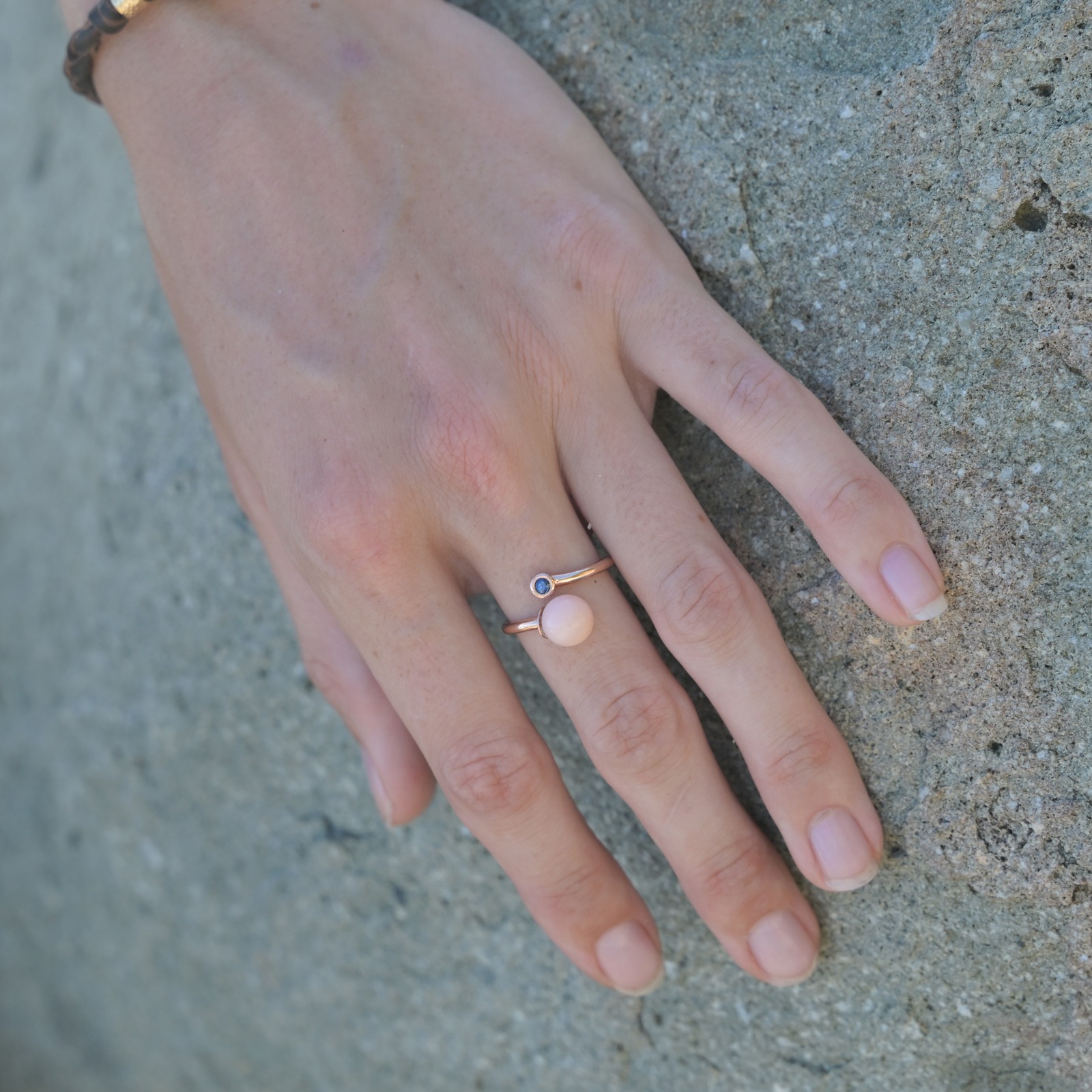 Bague Toi et Moi en or, perle de lambis et saphir, portée sur une main posée sur un rocher.