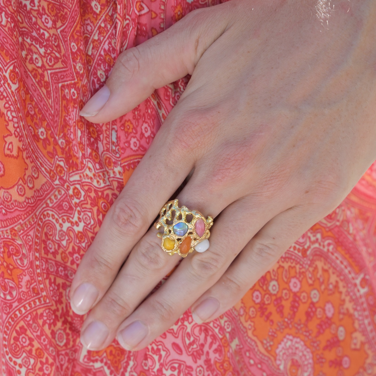 Bague Corail en or et perles fines multicolores, portée sur une main devant une robe rouge.