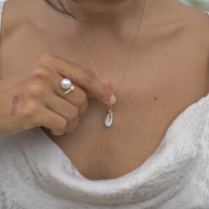 Gold pendant with an elongated natural clam pearl, worn on its chain, with one hand near the pearl.