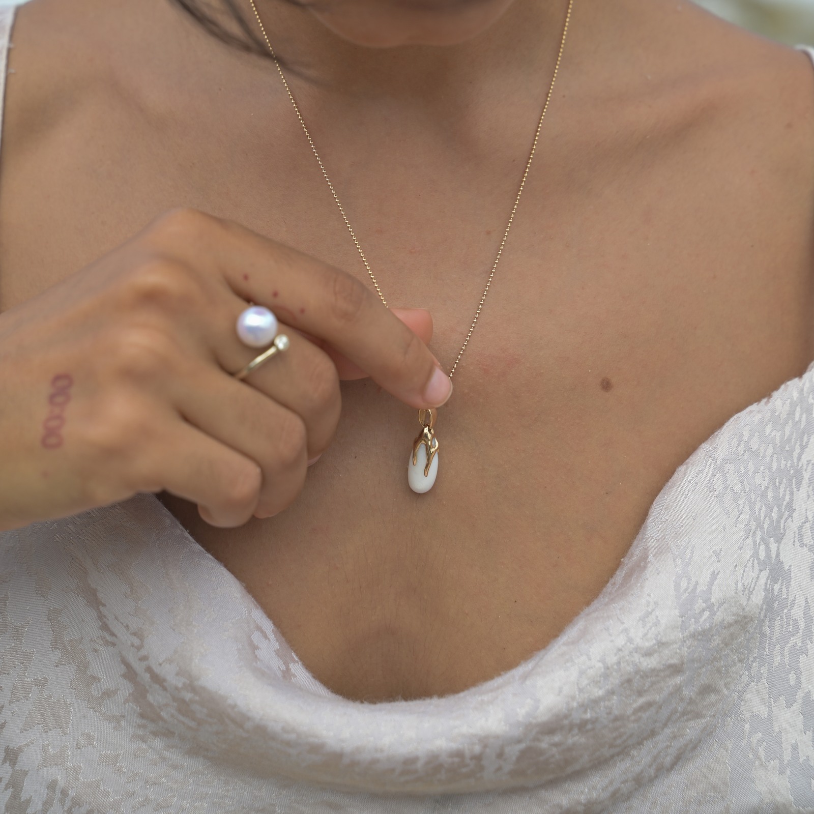 Gold pendant with an elongated natural clam pearl, worn on its chain, with one hand near the pearl.