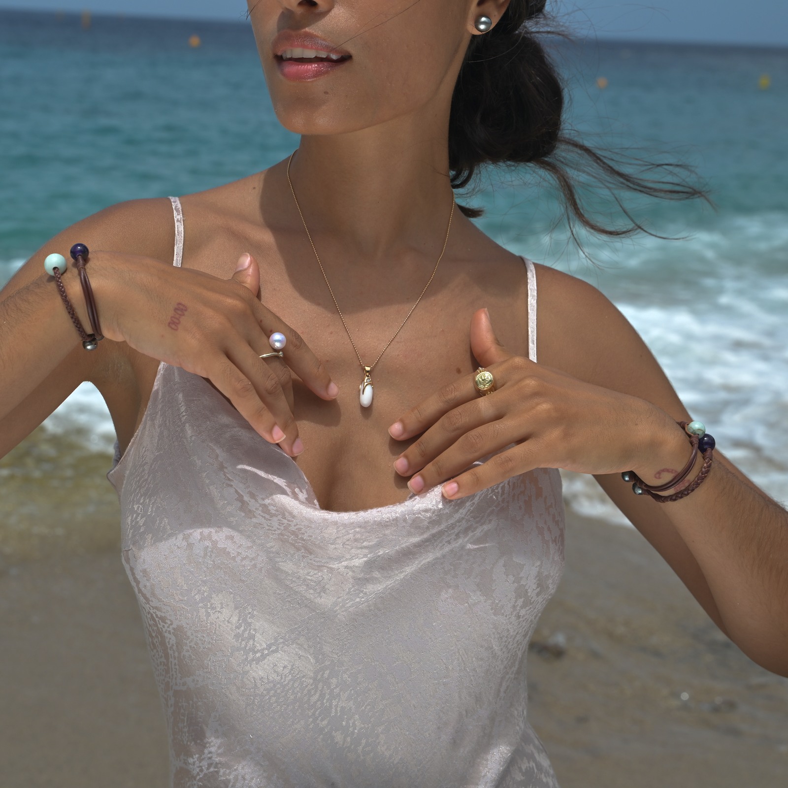 Gold pendant with an elongated natural clam pearl, worn by a model beside the sea, alongside other jewelry.