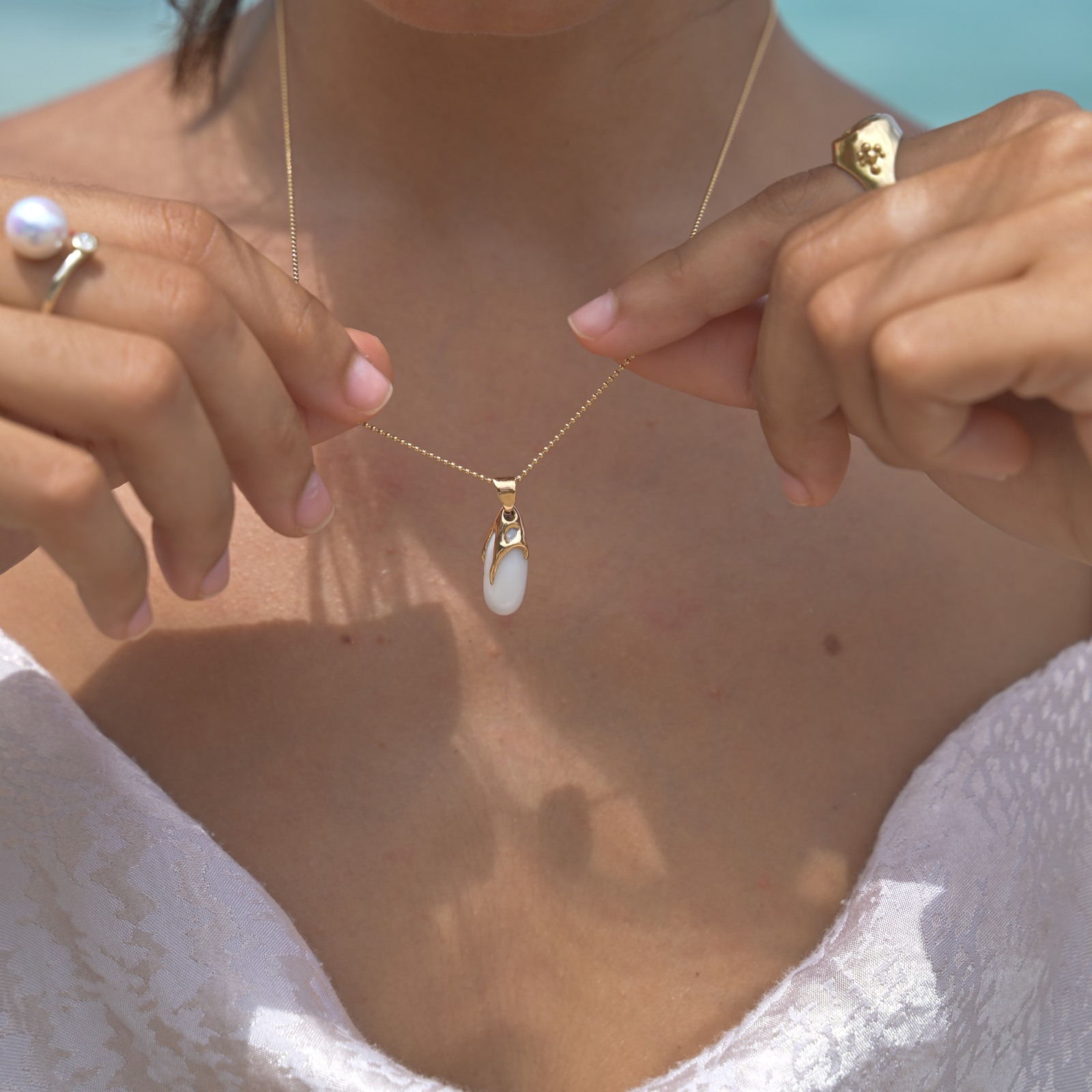 Gold pendant with an elongated natural clam pearl, worn with its chain held between both hands.